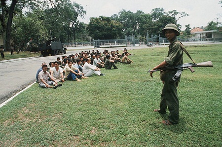 Những người làm việc cho chế độ cũ ngồi tập trung ở một góc sân Dinh Độc lập trước sự giám sát của một chiến sĩ giải phóng quân. (Ảnh: Jean-Claude Labbe/Gamma-Rapho)