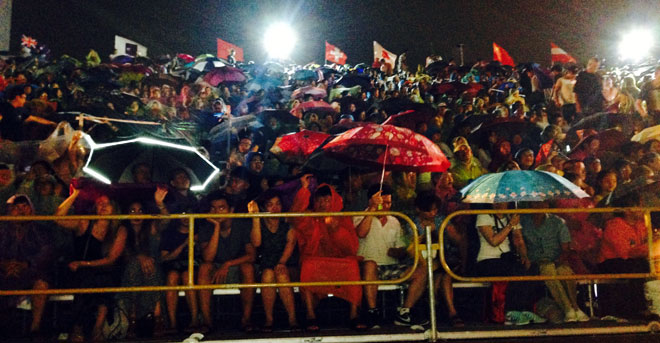 Locals and visitors wearing raincoats and holding umbrellas at viewing stands, …