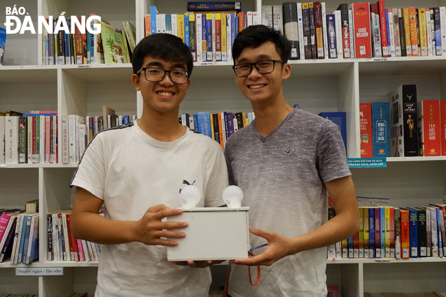 Nguyễn Trần Duy Khang (bên phải) và Nguyễn Minh Hiếu cùng sản phẩm công tắc nụ cười với thiết bị được kết nối là bóng đèn. Ảnh: MAI HIỀN