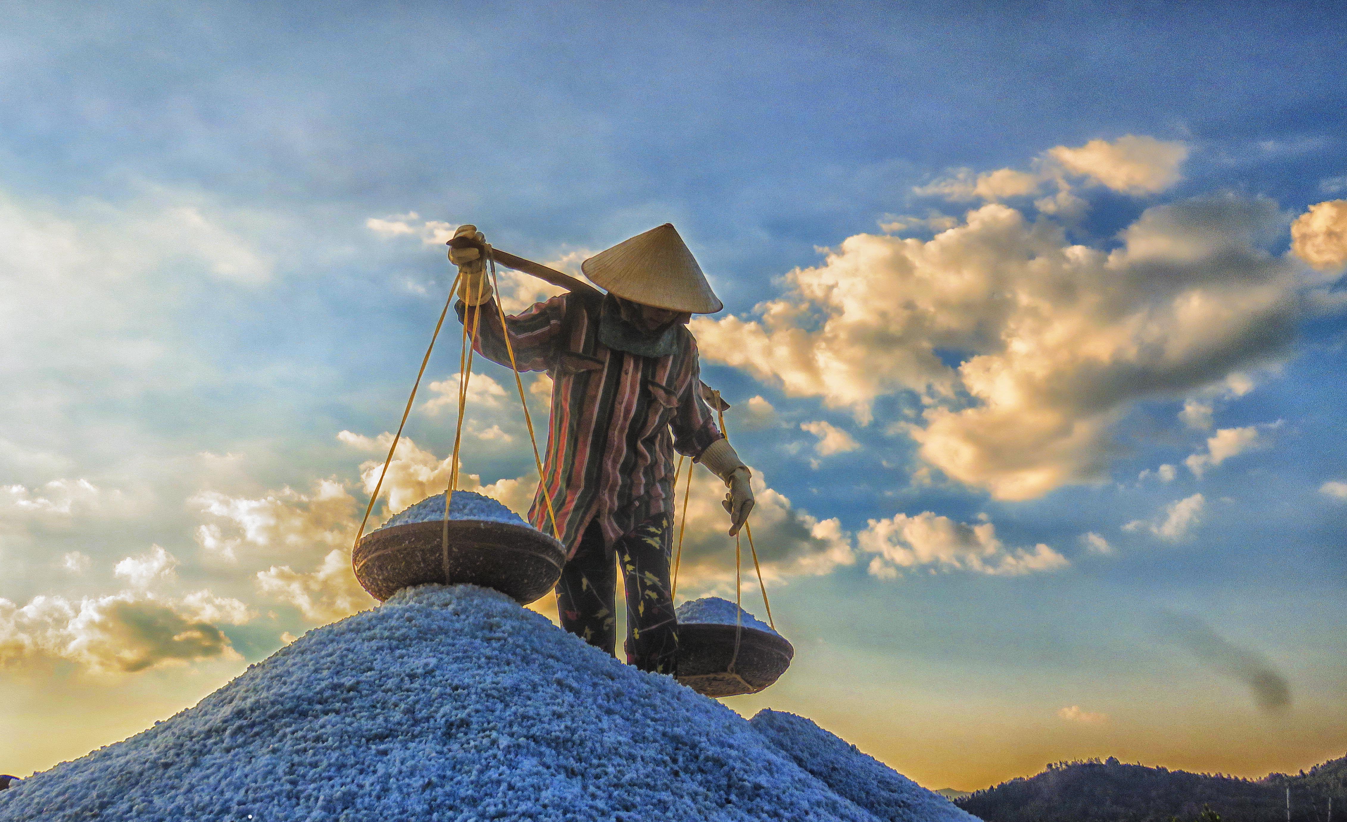 A female salt worker in a conical hat is seen deftly carrying two baskets of salt on bamboo poles slung over their shoulders.