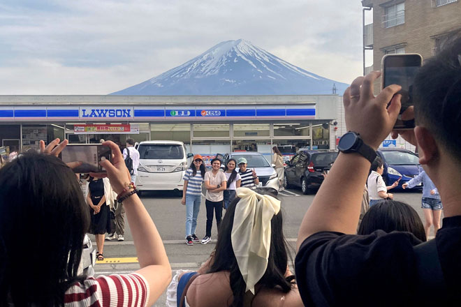Các du khách chụp hình kỷ niệm trước cửa hàng tiện lợi Lawson ở thị trấn Fujikawaguchiko, tỉnh Yamanashi, Nhật Bản với hình nền phía sau là núi Phú Sĩ vào ngày 28-4-2024.  Ảnh: Kyodo News / AP 